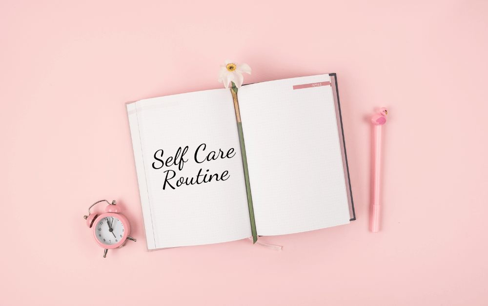 Flat lay of a self care journal, pink alarm clock, and pen on a pink background, emphasizing self care and a consistent daily routine.