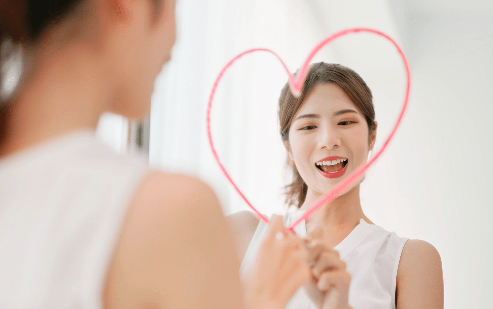 Woman smiling at her reflection after drawing a heart on the mirror with lipstick, embracing the message to be kind to yourself.