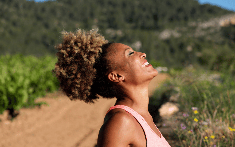 Smiling woman with curly hair enjoying nature outdoors, representing the mental health benefits of being outside and green therapy for emotional wellness.