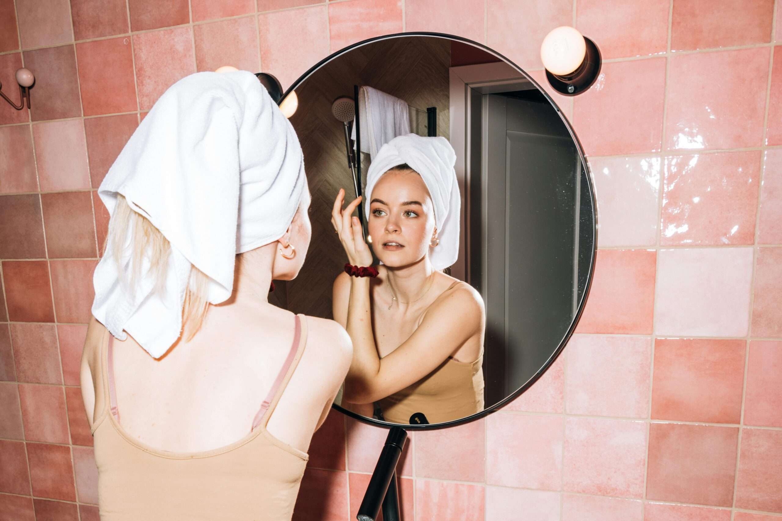 Woman enjoying her beauty routine with self-care products, wearing a towel on her head, and applying skincare in a calm, relaxing environment.