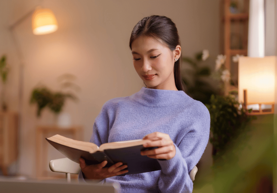 A smiling woman reading a book in a cozy space, representing how to better yourself through personal improvement, self-love, and daily self-improvement habits for personal growth.